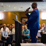 Patrick Vernon on stage holding a microphone as he addresses the audience of the conference